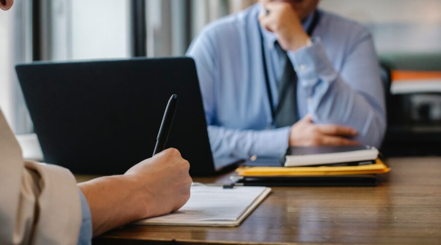 Twee personen tijdens een zakelijk gesprek aan een bureau, waarbij één persoon schrijft op een notitieblok en de ander luistert, met een laptop en documenten op tafel.